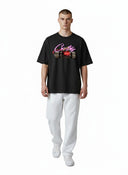 Hombre vistiendo camiseta negra Corteiz No Time 4 Luv con estampado de dinamita rosa, de One Kickz